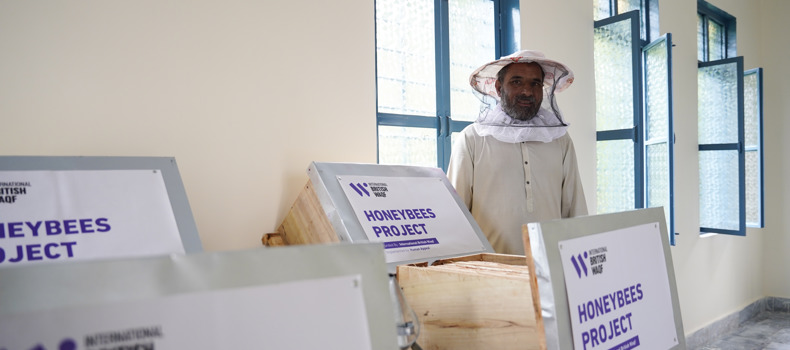 فوائد الوقف الخيري, شخص يرتدي بدلة تربية النحل يقف في غرفة تحتوي على عدة صناديق لتربية النحل تحمل علامة "مشروع النحل" من قبل الوقف البريطاني الدولي. تحتوي الغرفة على نوافذ متعددة بإطارات زرقاء ومصاريع مفتوحة، مما يسمح بدخول الضوء الطبيعي.
