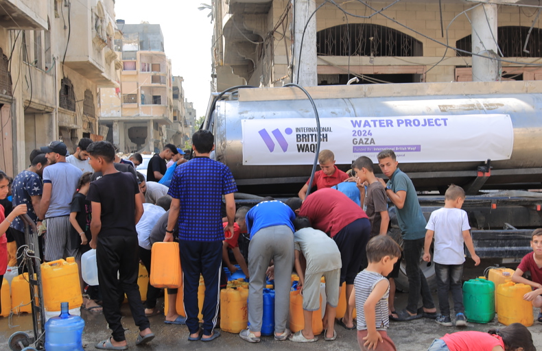 ما هو الوقف الخيري مجموعة من الأشخاص، بينهم أطفال وشباب، يتجمعون حول صهريج ماء في منطقة متضررة، ويملؤون عبوات بلاستيكية بالمياه. اللافتة على الصهريج تشير إلى مشروع مياه 2024 في غزة، بتمويل من منظمة الوقف البريطاني الدولي
